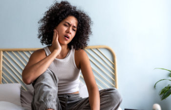 young woman with curly hair touching cheek in pain having toothache sitting on bed, 