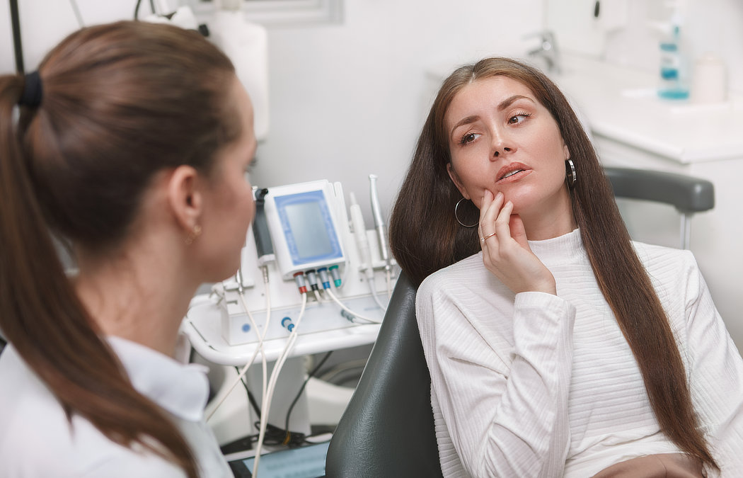 A woman in a dentist's chair touches her face, looking concerned, while a dental professional listens. Dental equipment is visible in the background., 