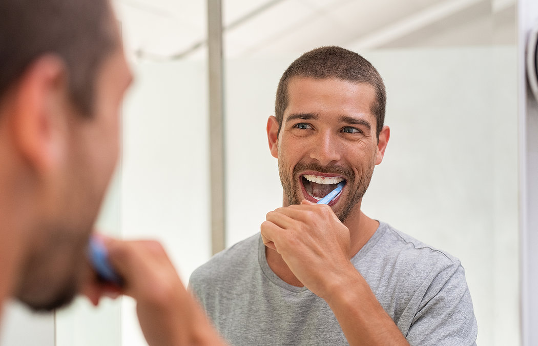 A man brushing his teeth in front of a bathroom mirror, wearing a gray t-shirt., 