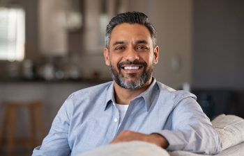 Smiling man with short hair and beard, wearing a light blue shirt, sitting on a sofa in a cozy living room.